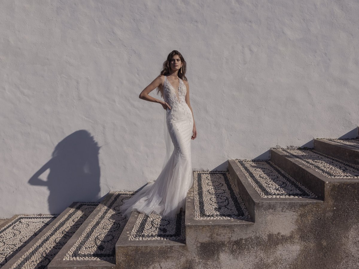 Woman in an elegant white lace wedding dress with flowing train standing on geometric stone steps against a white wall