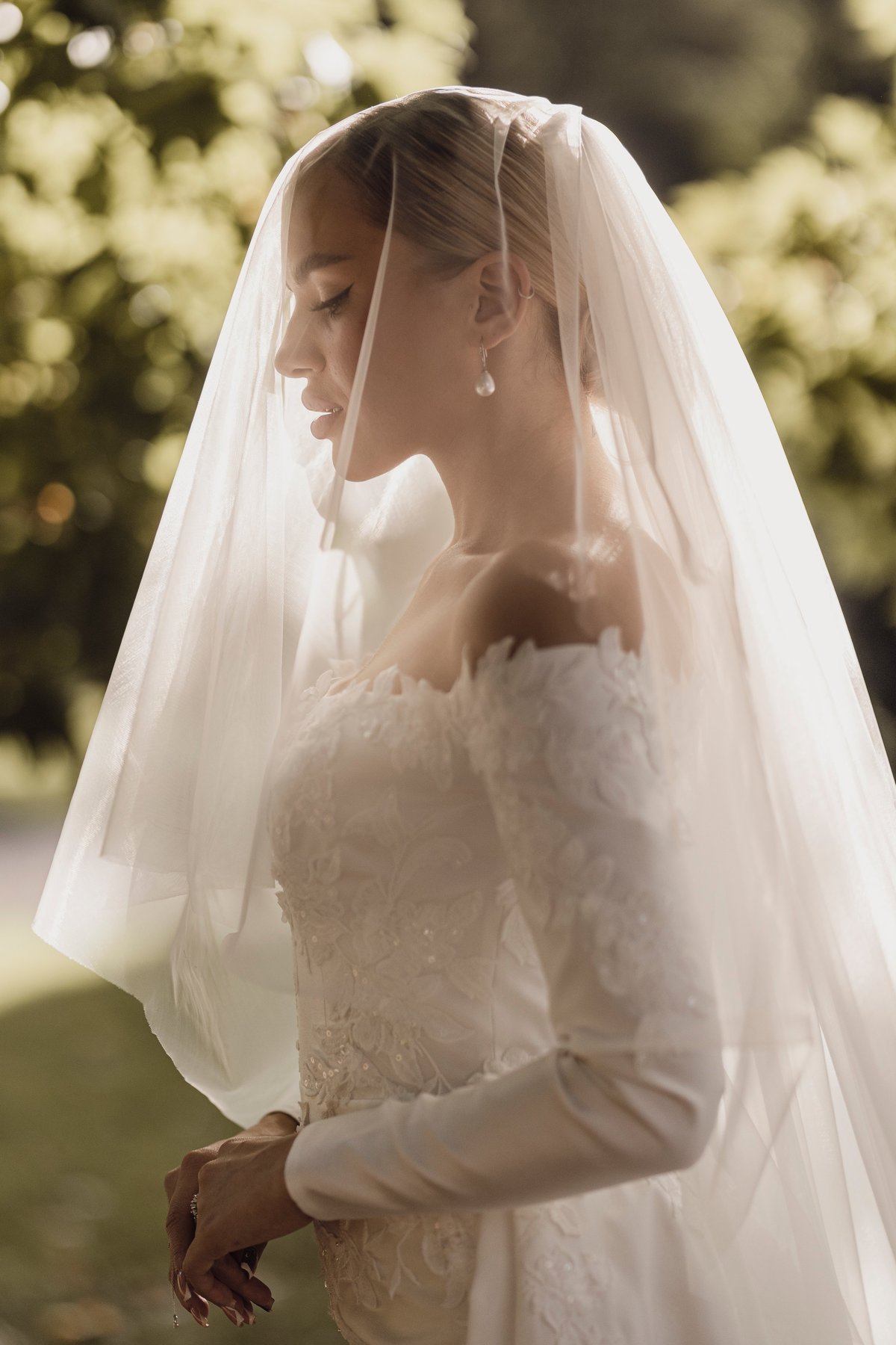 Bride in white lace wedding dress with long veil standing outdoors in soft natural light