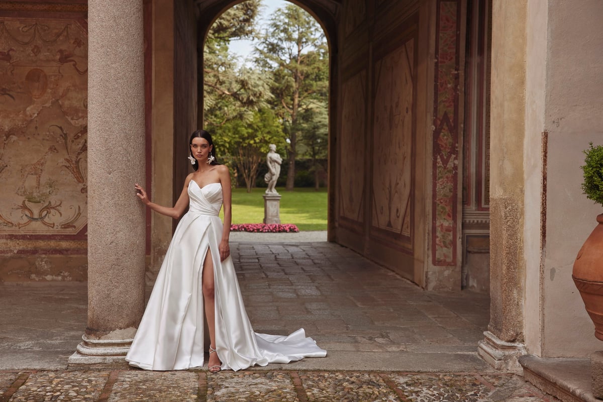Bride in white wedding dress with high slit standing in arched colonnade, garden visible through doorway