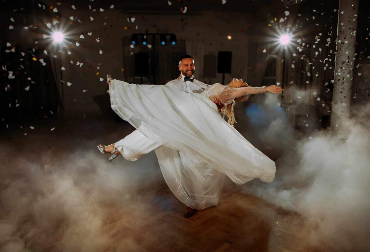 Groom in tuxedo twirling bride in white wedding dress during first dance with dramatic fog and lighting effects