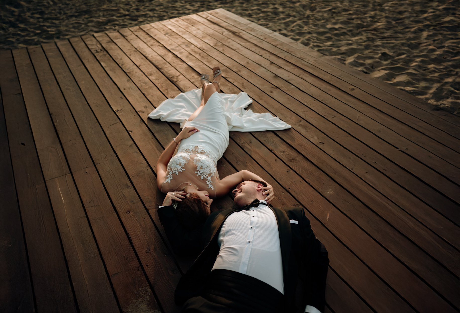 Person lying on a wooden boardwalk by the beach, wearing casual clothing, viewed from above.