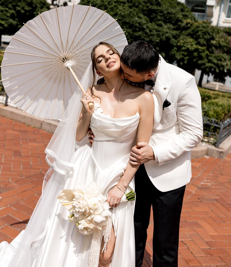 Bride and groom on brick patio. Bride wears white dress and holds umbrella, groom wears light blazer. Bride holds white flower bouquet