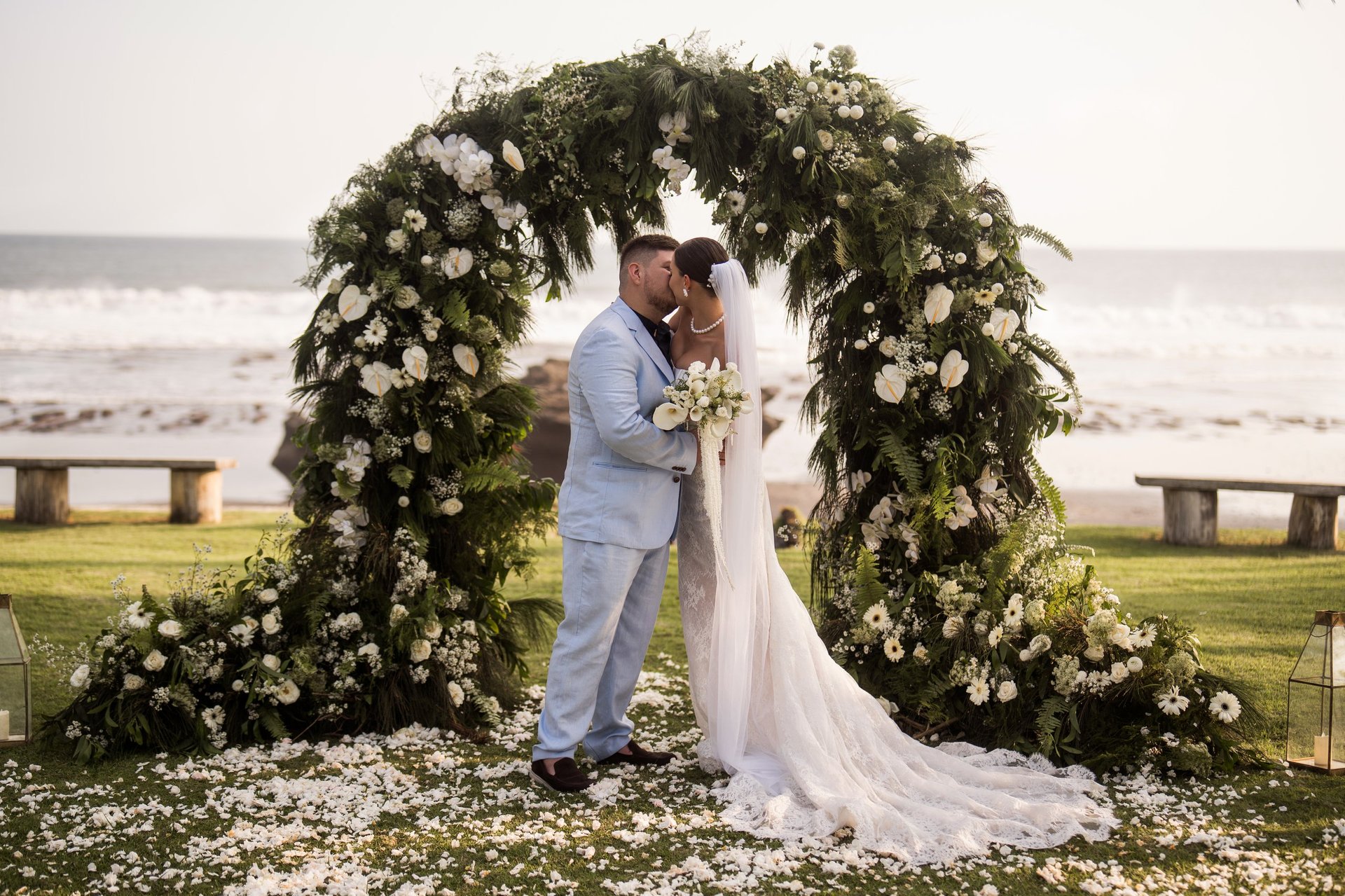 Bride and groom kissing under a floral arch on a beach with ocean waves in the background