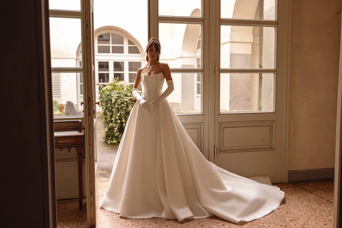 Bride in elegant white wedding gown with long train standing in doorway of classic beige room with tall windows