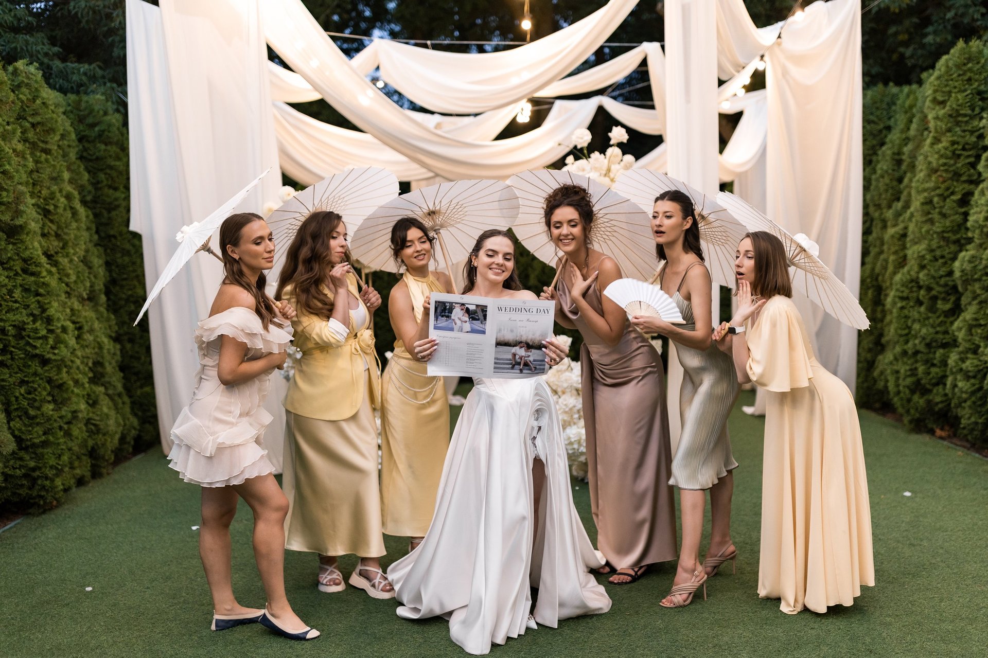 Bride and bridesmaids in elegant gowns standing together under draped white fabric canopy holding photos outdoors