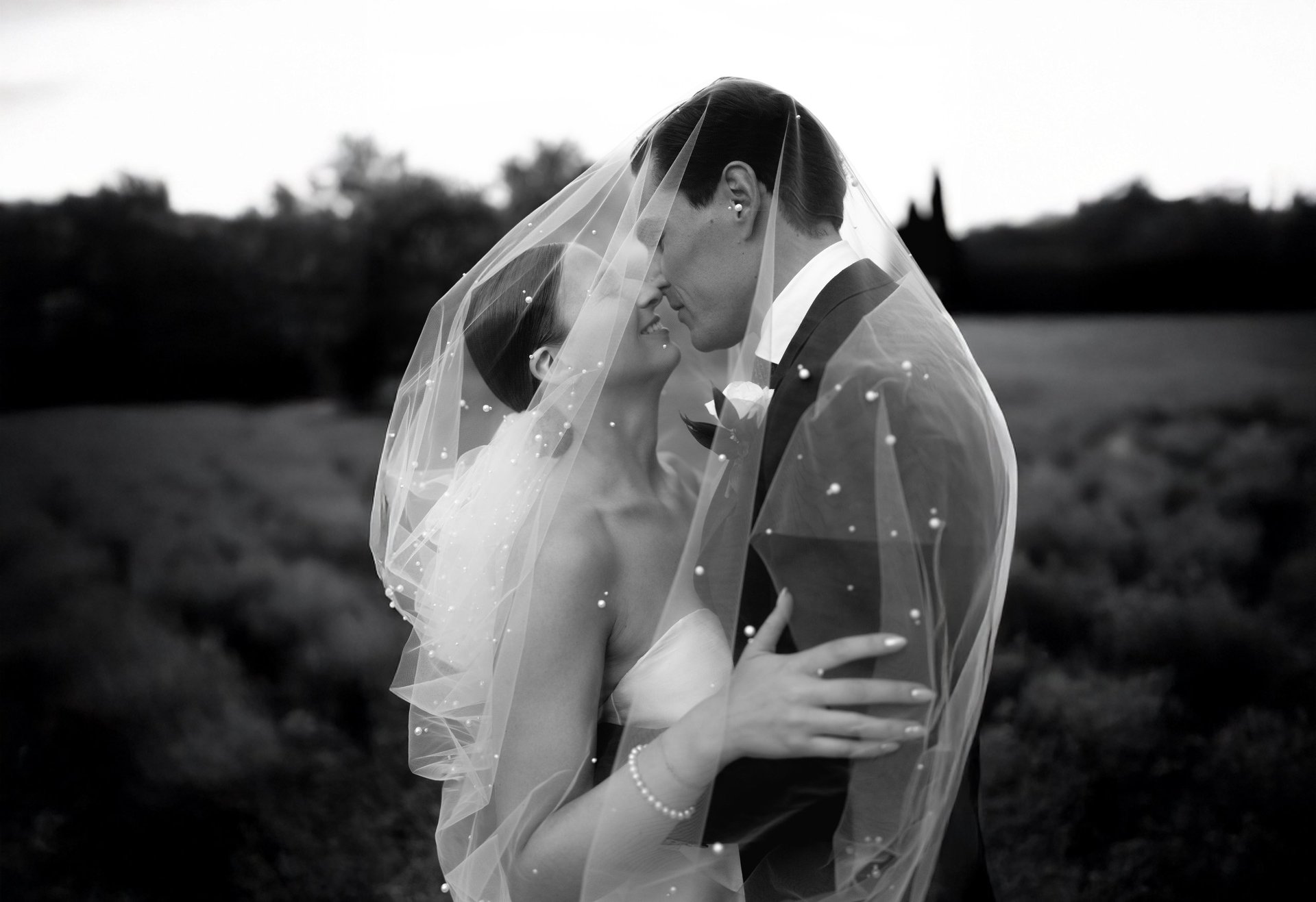 Black and white wedding photo of bride and groom embracing under bridal veil in outdoor field setting