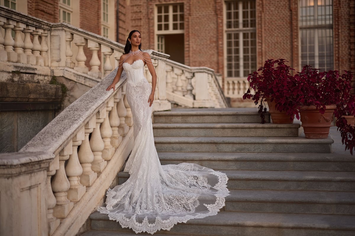 Bride in white lace gown with long train posing on grand stone staircase beside ornate balustrade and terracotta planter