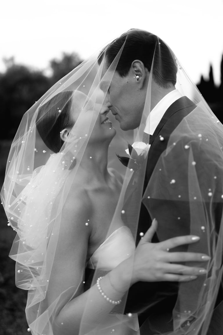 Black and white photo of bride and groom kissing under beaded wedding veil outdoors with blurred trees in background
